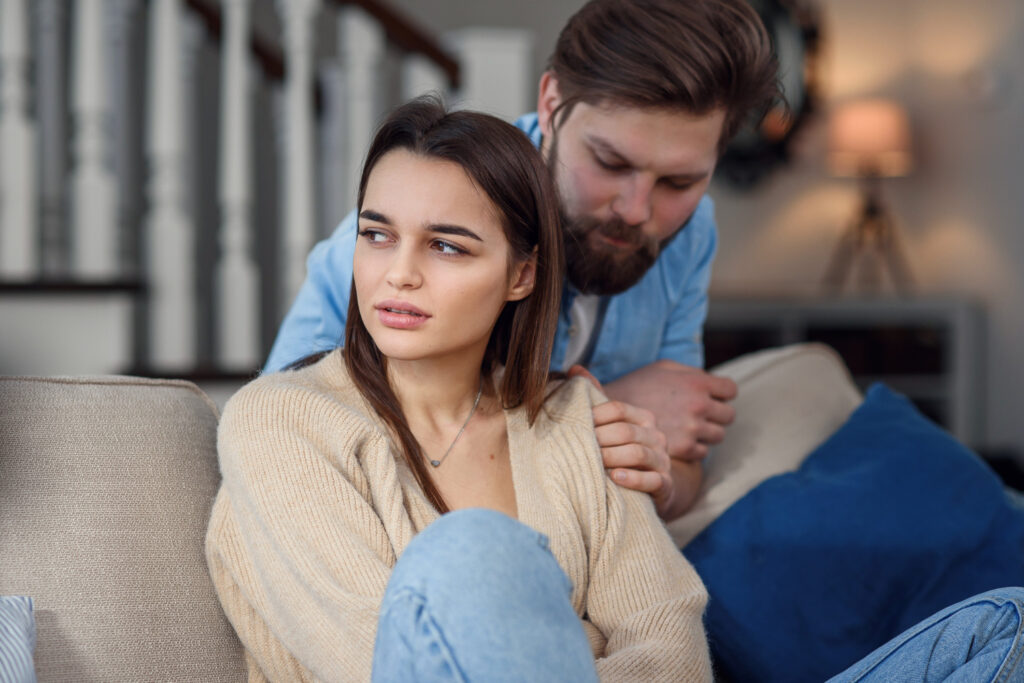 Brodaty mężczyzna w niebieskiej koszuli próbuje rozmawiać z partnerką, siedzącą przed nim na kanapie i odwracającą od niego głowę.