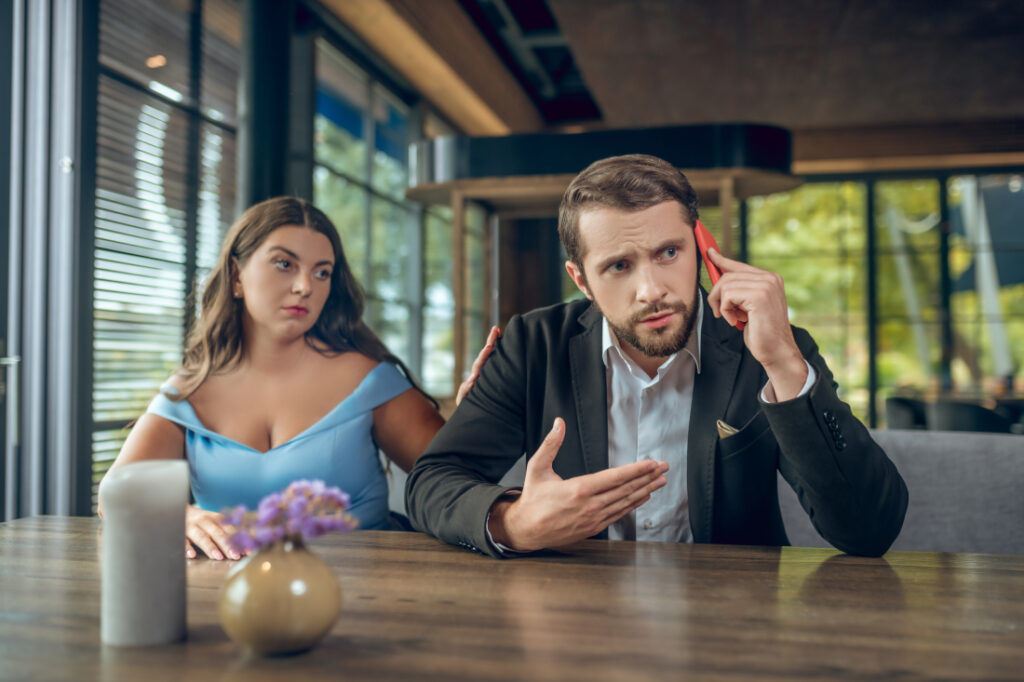 Mężczyzna ubrany w garnitur w trakcie randki w restauracji z kobietą w błękitnej sukience rozmawia rozzłoszczony przez telefon komórkowy